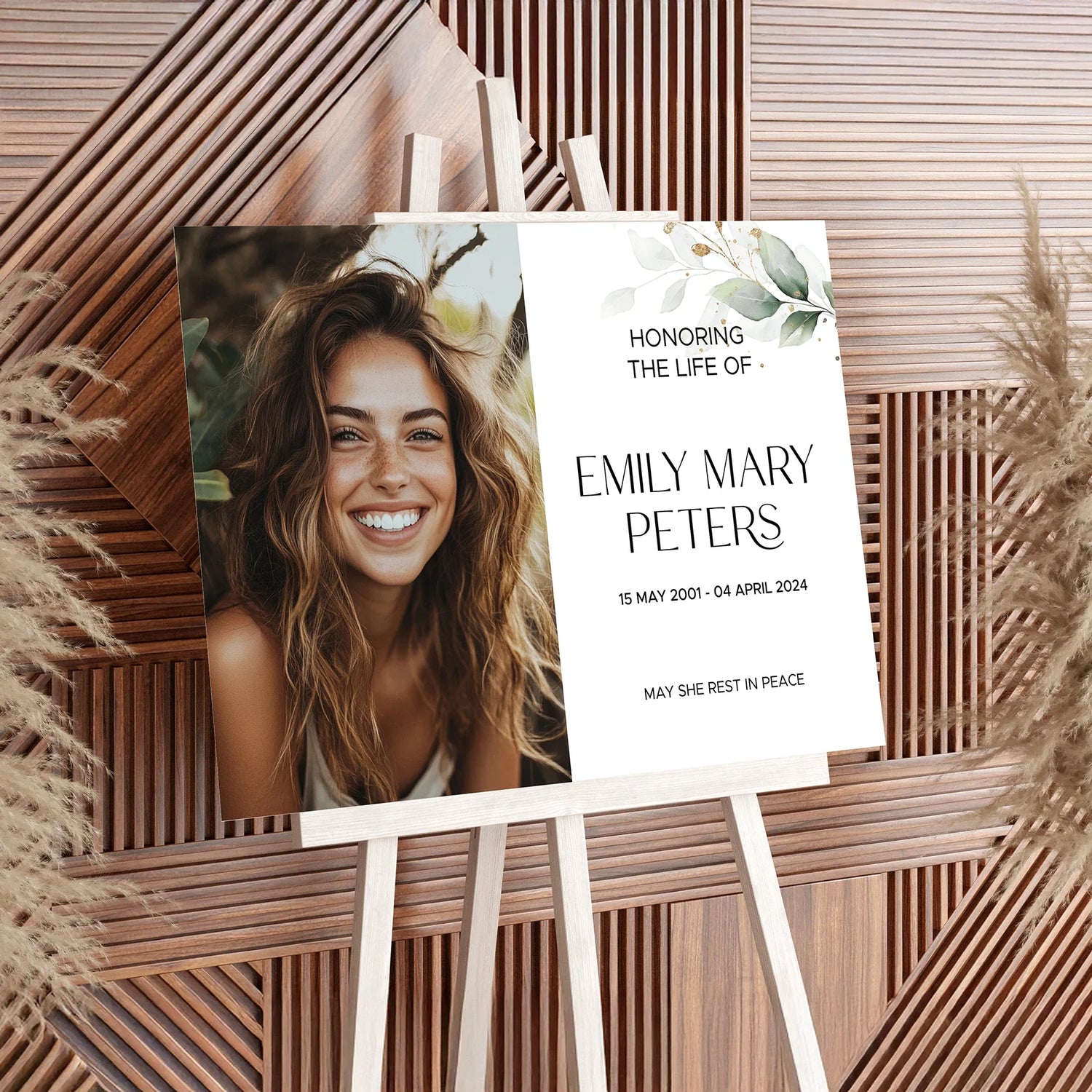 Memorial welcome sign on an easel, featuring a smiling portrait of Emily Mary Peters, floral greenery accents, and text honoring her life with birth and passing dates. A beautiful tribute displayed against a wooden backdrop for a remembrance service.