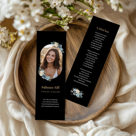 Memorial bookmark for Katherine Hill (2021–2024) are displayed on a wooden plate with white flowers around. One features her portrait with a remembrance quote, while the other contains the poem "A Silent Tear." Both sides have a black background with elegant blue and white floral accents.