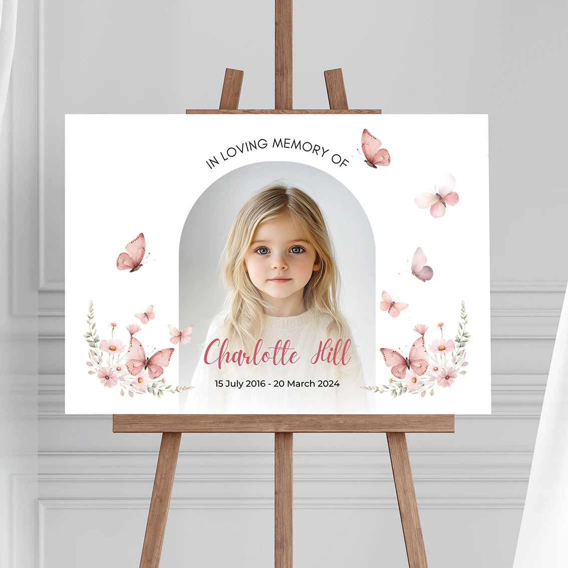 Memorial sign on a wooden easel at the beach, honoring a young girl named Charlotte Hill. Features a soft floral and butterfly design, her photo, name, and dates. Surrounded by white roses, candles, and flowers, creating a peaceful tribute.