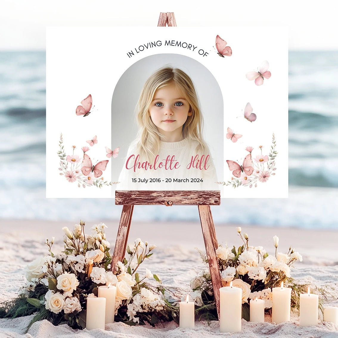 Memorial sign on a wooden easel at the beach, honoring a young girl named Charlotte Hill. Features a soft floral and butterfly design, her photo, name, and dates. Surrounded by white roses, candles, and flowers, creating a peaceful tribute.