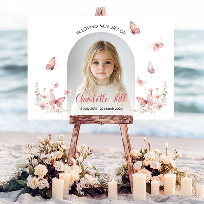 Memorial sign on a wooden easel at the beach, honoring a young girl named Charlotte Hill. Features a soft floral and butterfly design, her photo, name, and dates. Surrounded by white roses, candles, and flowers, creating a peaceful tribute.