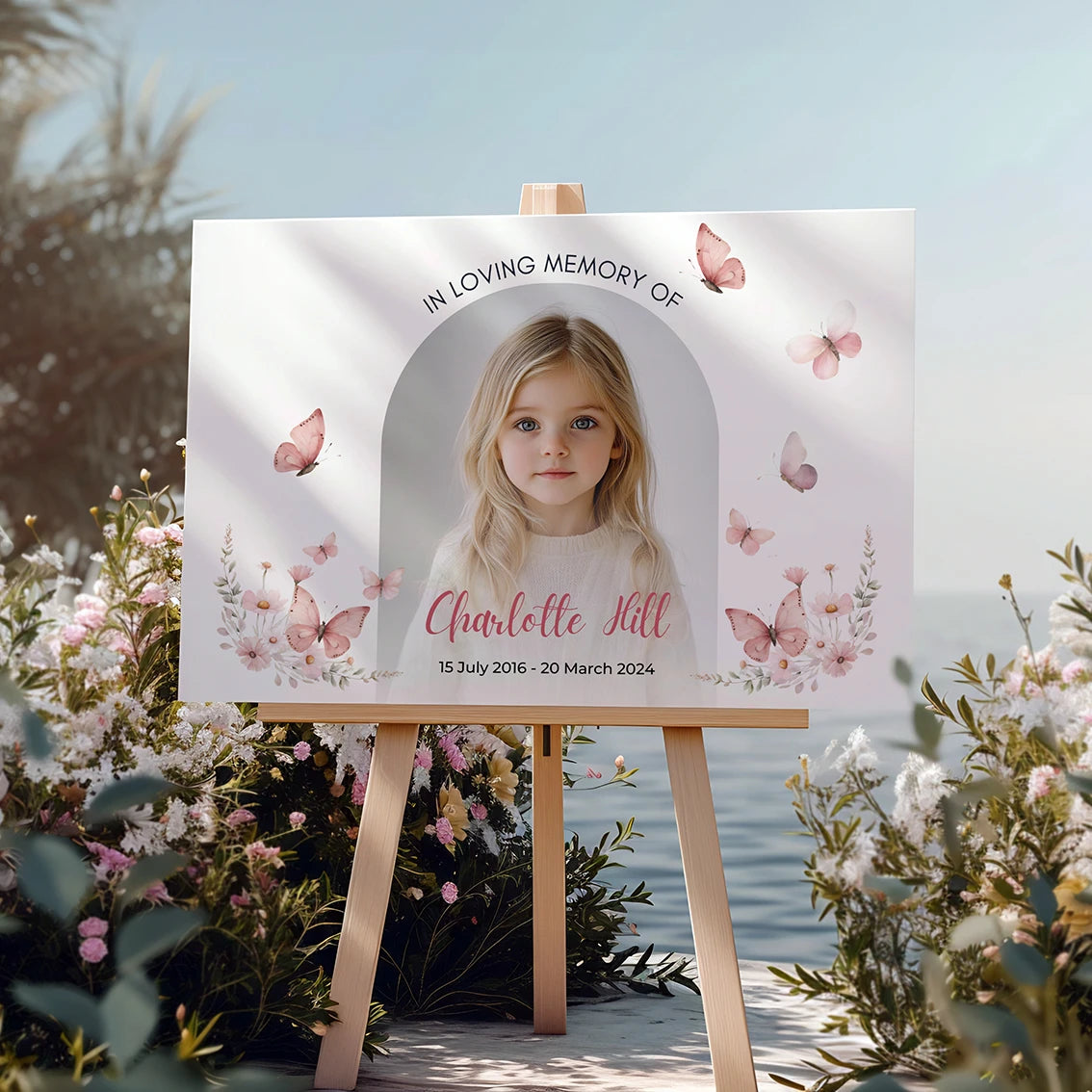 Memorial sign on a wooden easel at the beach, honoring a young girl named Charlotte Hill. Features a soft floral and butterfly design, her photo, name, and dates. Surrounded by white roses, candles, and flowers, creating a peaceful tribute.