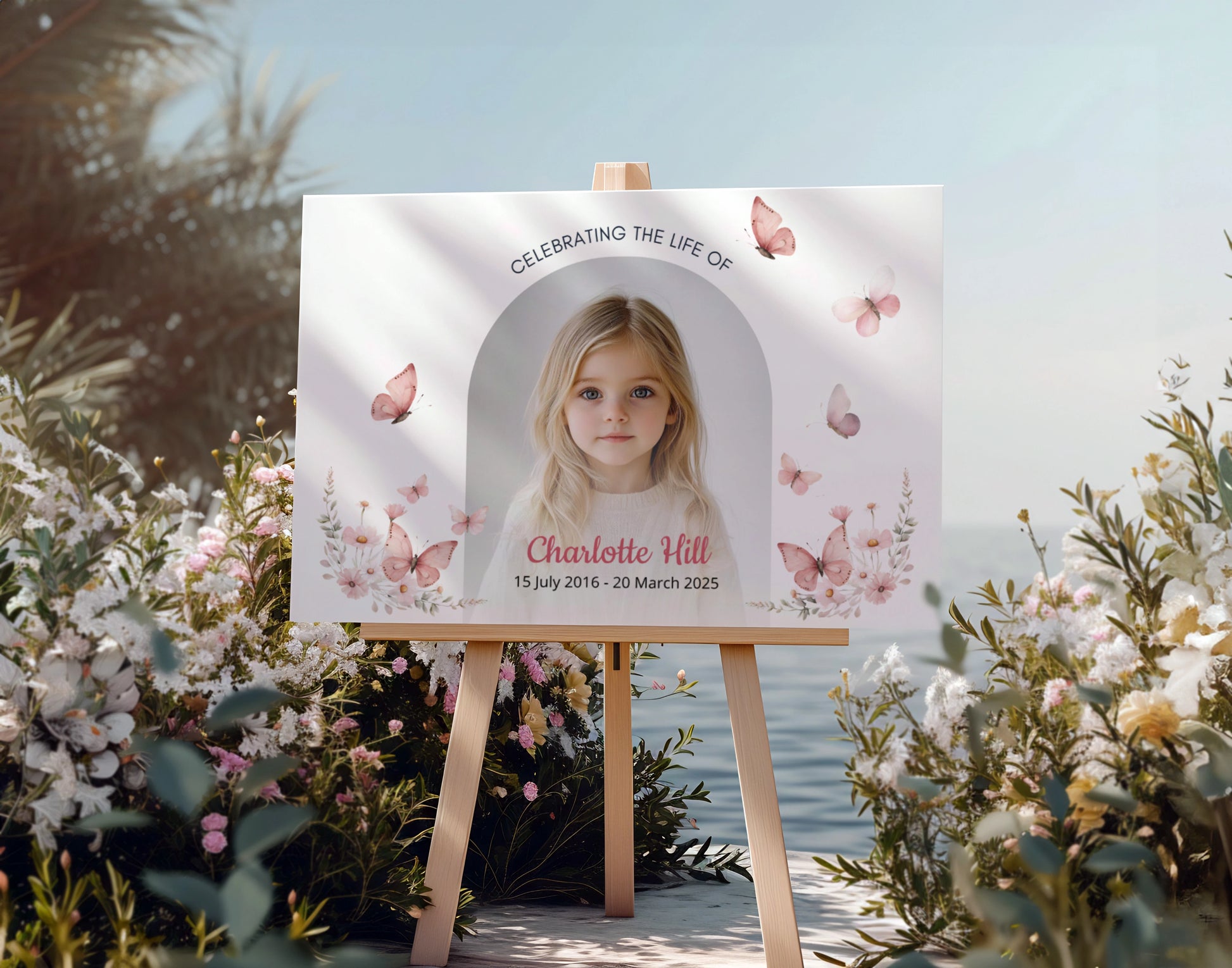 Memorial sign on a wooden easel at the beach, honoring a young girl named Charlotte Hill. Features a soft floral and butterfly design, her photo, name, and dates. Surrounded by white roses, candles, and flowers, creating a peaceful tribute.