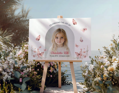 Memorial sign on a wooden easel at the beach, honoring a young girl named Charlotte Hill. Features a soft floral and butterfly design, her photo, name, and dates. Surrounded by white roses, candles, and flowers, creating a peaceful tribute.
