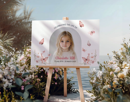 Memorial sign on a wooden easel at the beach, honoring a young girl named Charlotte Hill. Features a soft floral and butterfly design, her photo, name, and dates. Surrounded by white roses, candles, and flowers, creating a peaceful tribute.