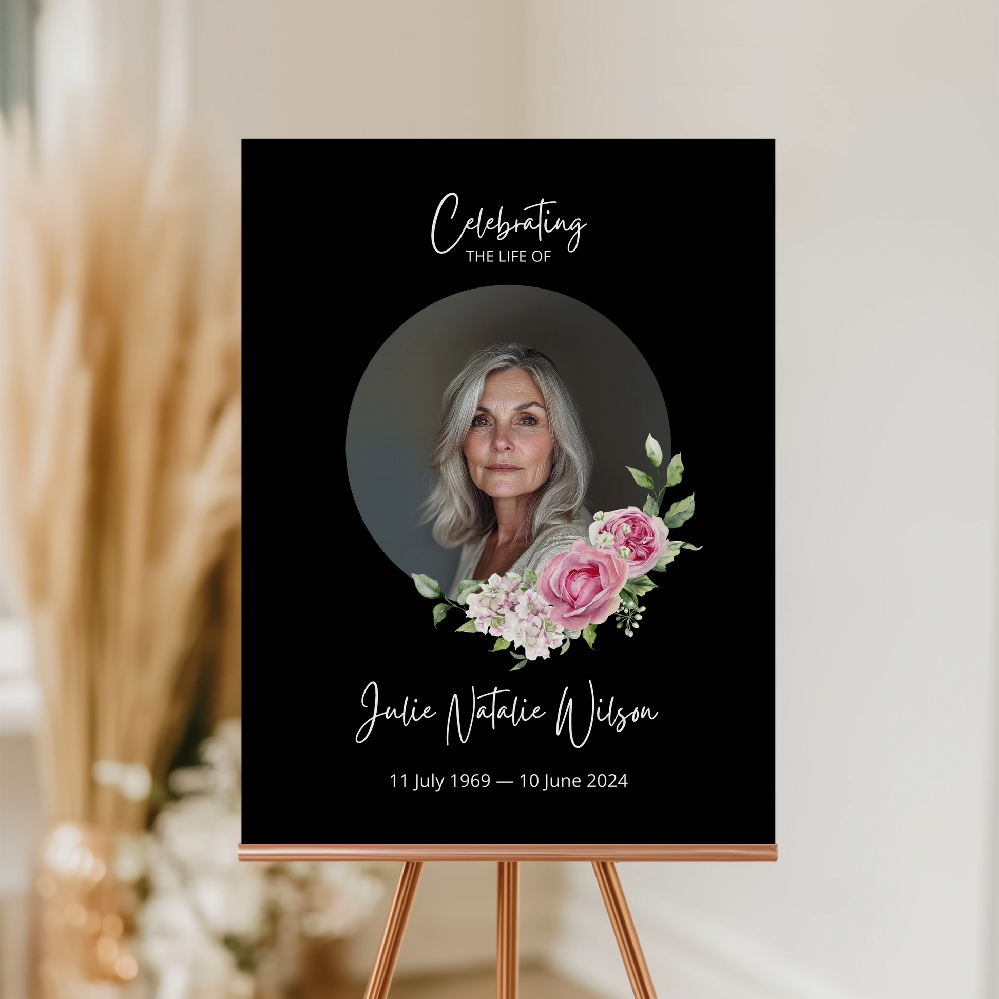 Memorial sign with a black background, featuring a circular portrait of a woman and pink floral accents. The text honours "Julie Natalie Wilson" with her birth and passing dates, serving as a beautiful tribute.