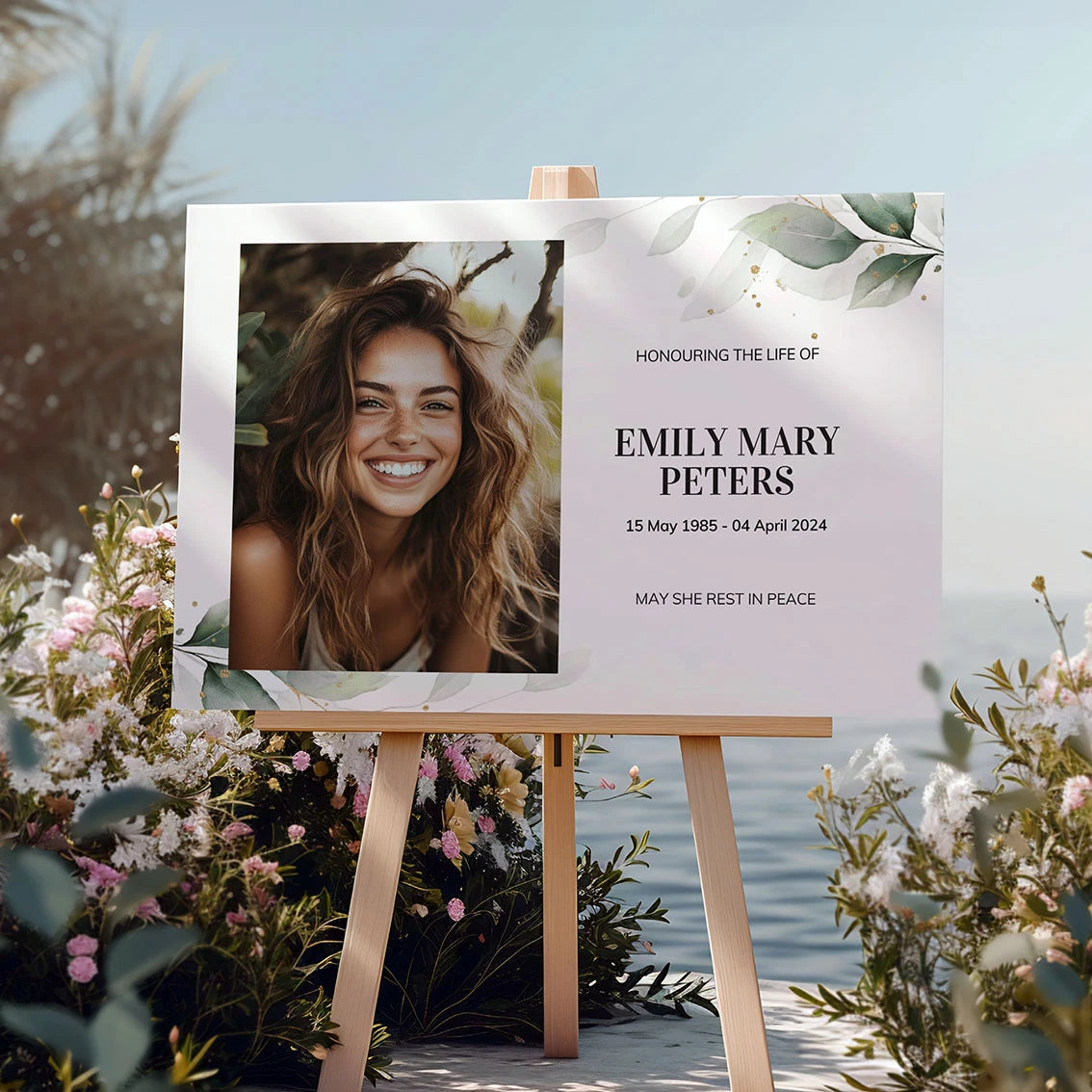 Memorial sign on a wooden easel at the beach, honouring a woman named Mary Louise Wilson. Features a soft eucalyptus design, her photo, name, and dates. Surrounded by white roses, candles, and flowers, creating a peaceful tribute.
