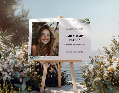 Memorial sign on a wooden easel at the beach, honouring a woman named Mary Louise Wilson. Features a soft eucalyptus design, her photo, name, and dates. Surrounded by white roses, candles, and flowers, creating a peaceful tribute.