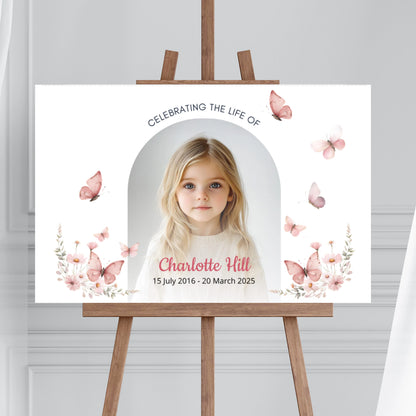 Memorial sign on a wooden easel at the beach, honouring a young girl named Charlotte Hill. Features a soft floral and butterfly design, her photo, name, and dates. Surrounded by white roses, candles, and flowers, creating a peaceful tribute.