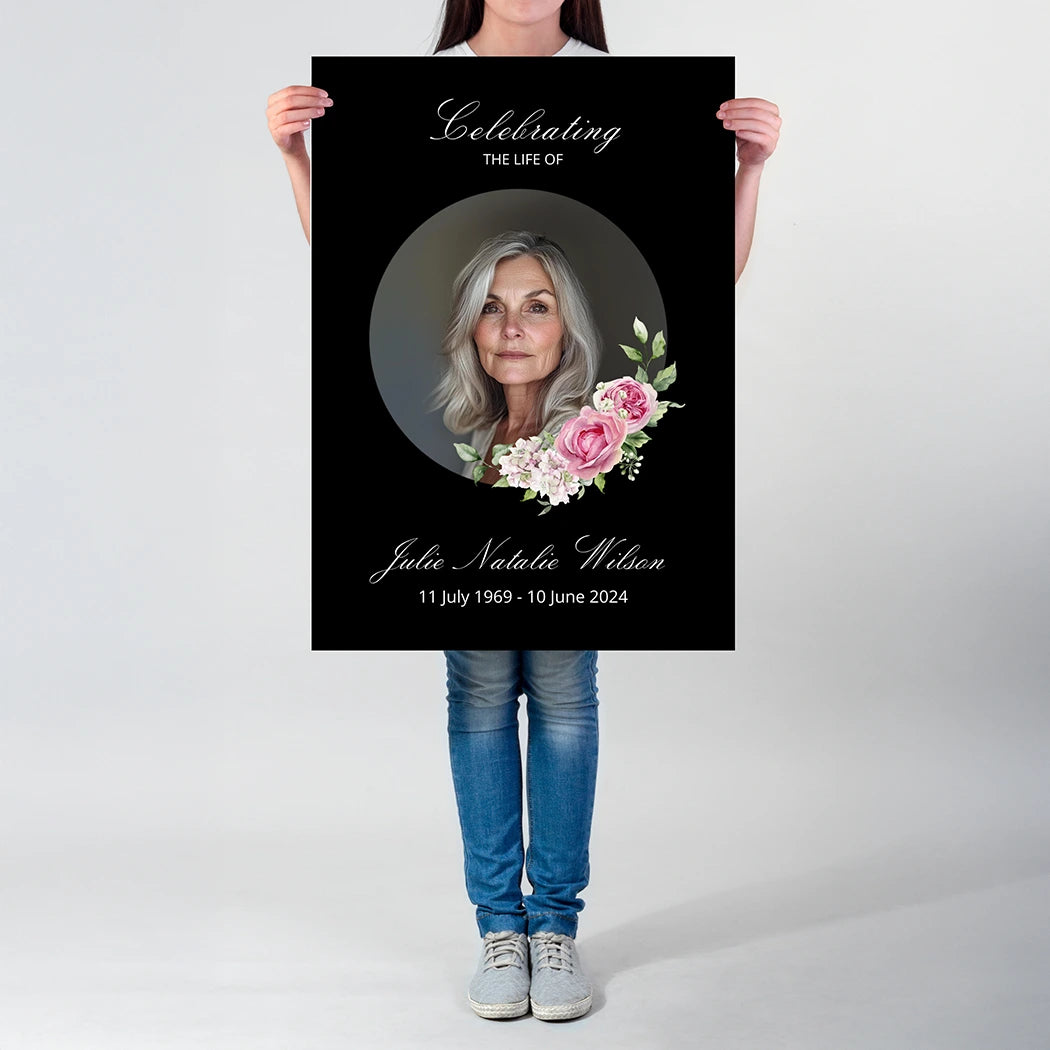 A person holding a memorial poster featuring a portrait, floral accents, and the person’s name written in cursive on a black background.