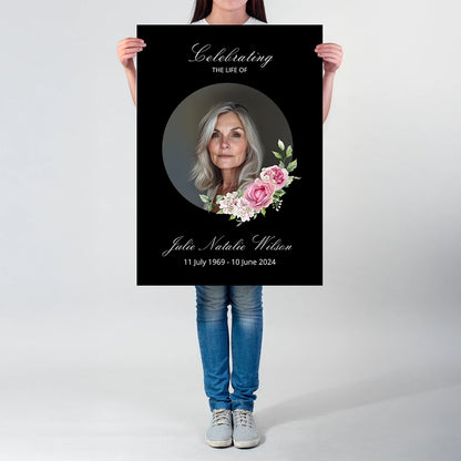 A person holding a memorial poster featuring a portrait, floral accents, and the person’s name written in cursive on a black background.